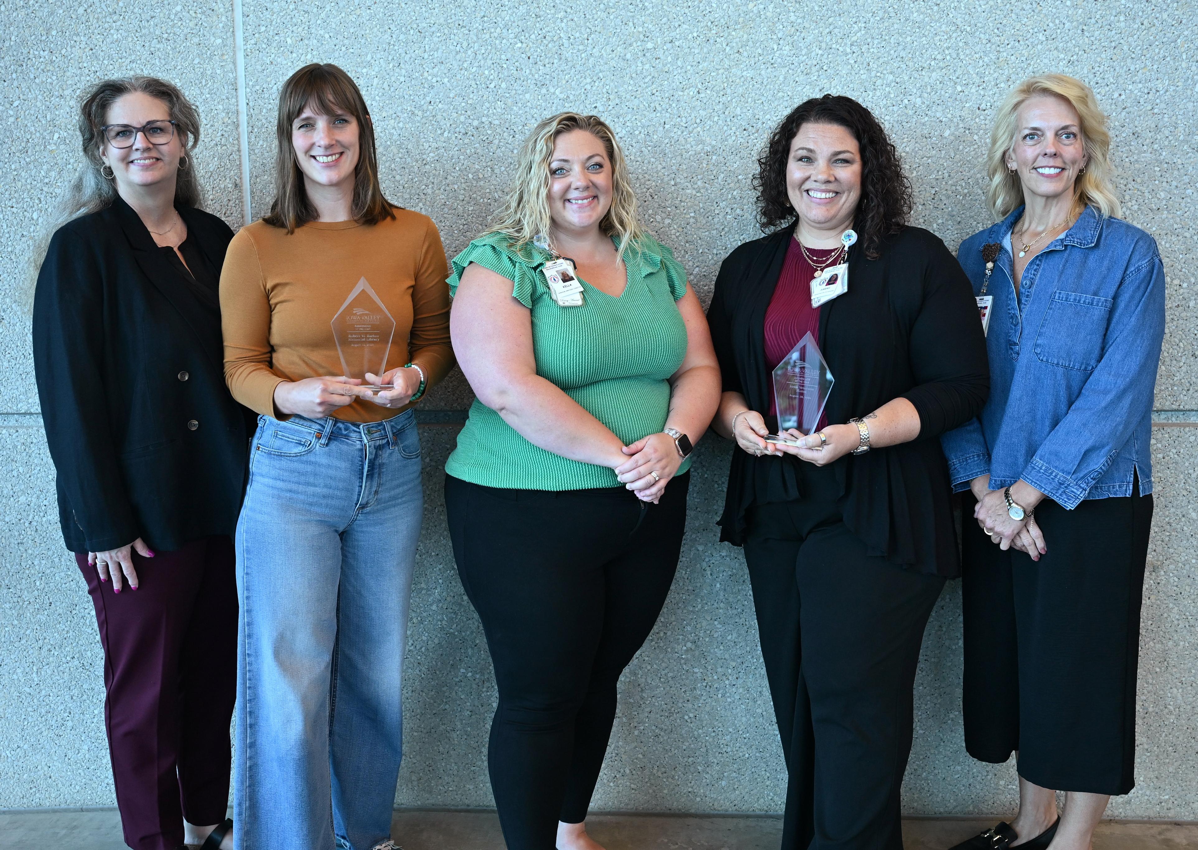 Five people stand in front of a textured indoor wall, with two individuals holding clear glass award plaques while the group poses for a recognition photo.