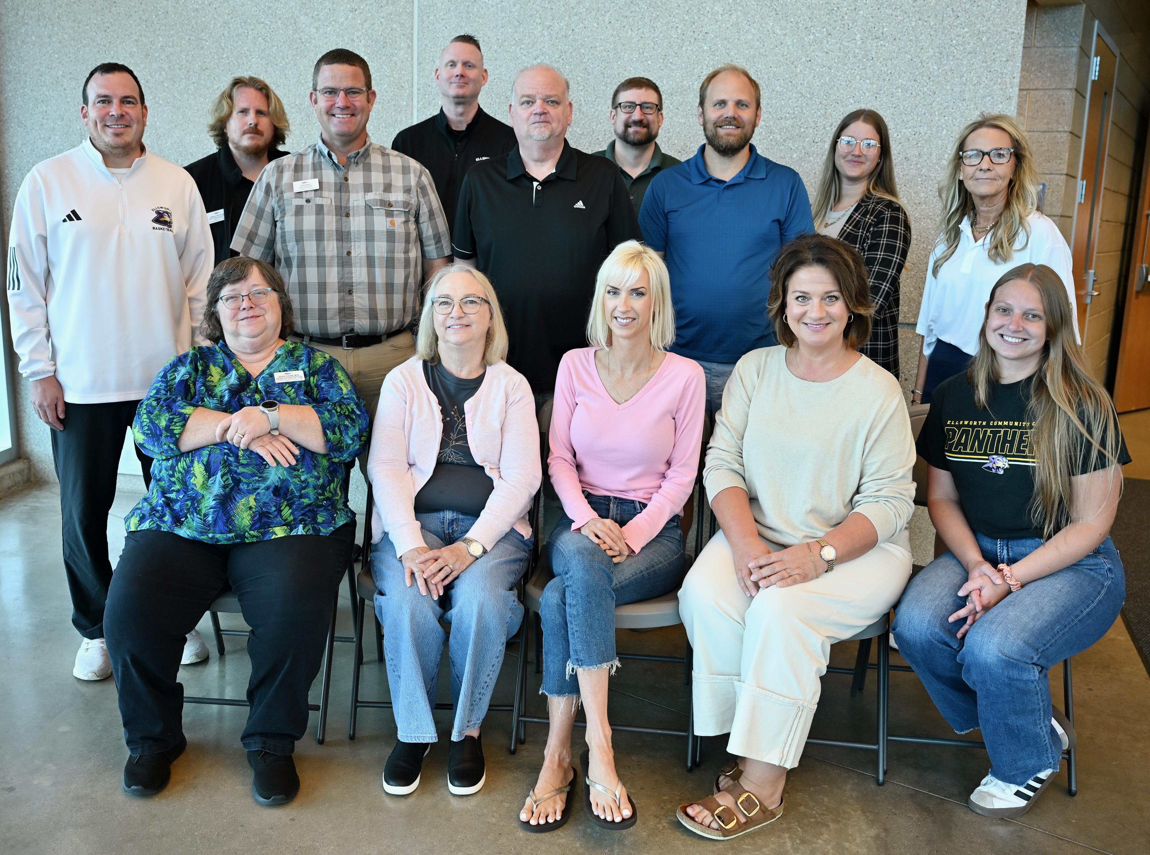 Group portrait of fourteen employees in an indoor facility, with five seated in the front row and nine standing behind them against a textured wall.