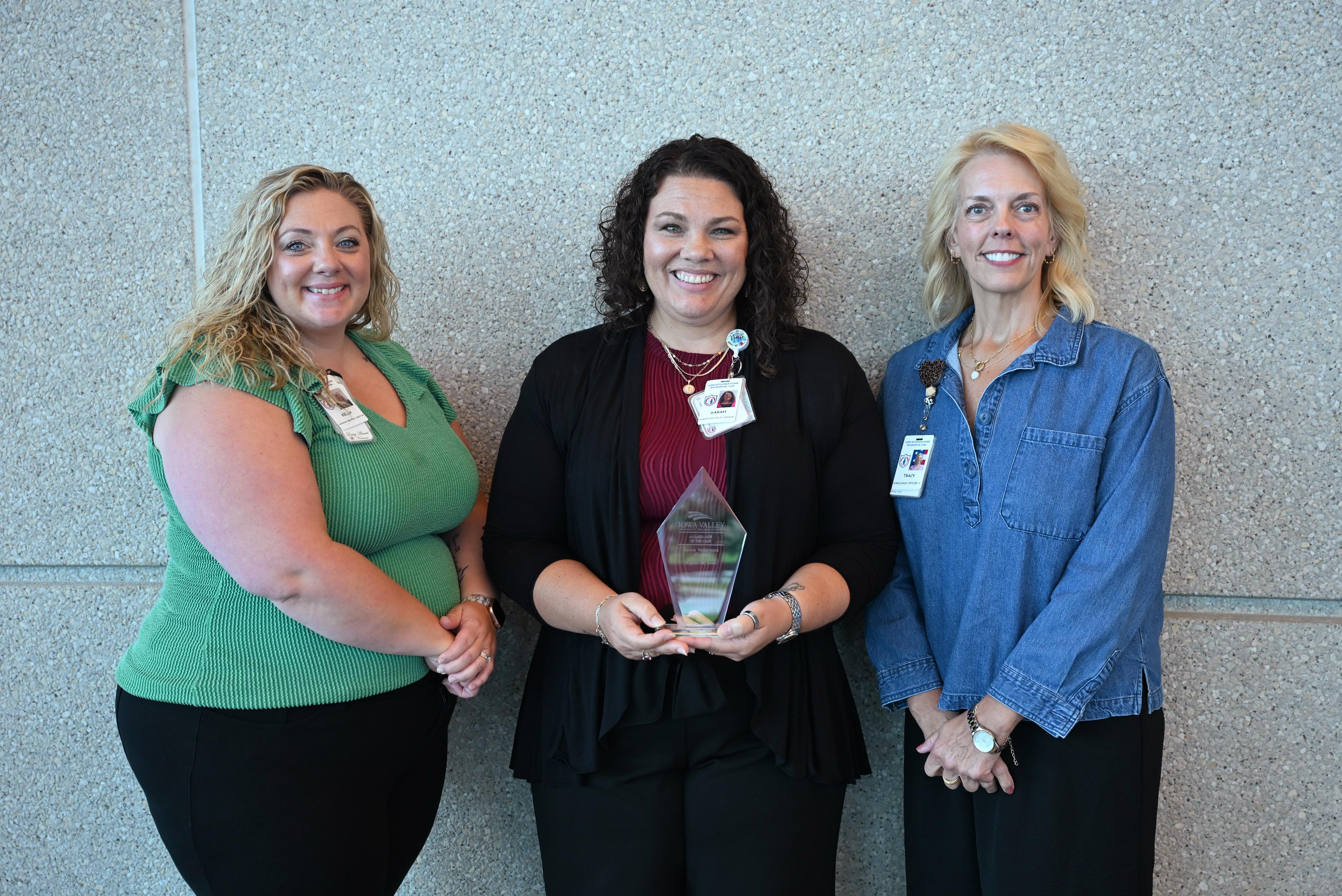 Three people pose for a recognition photo, with the person in the center holding a glass award plaque.