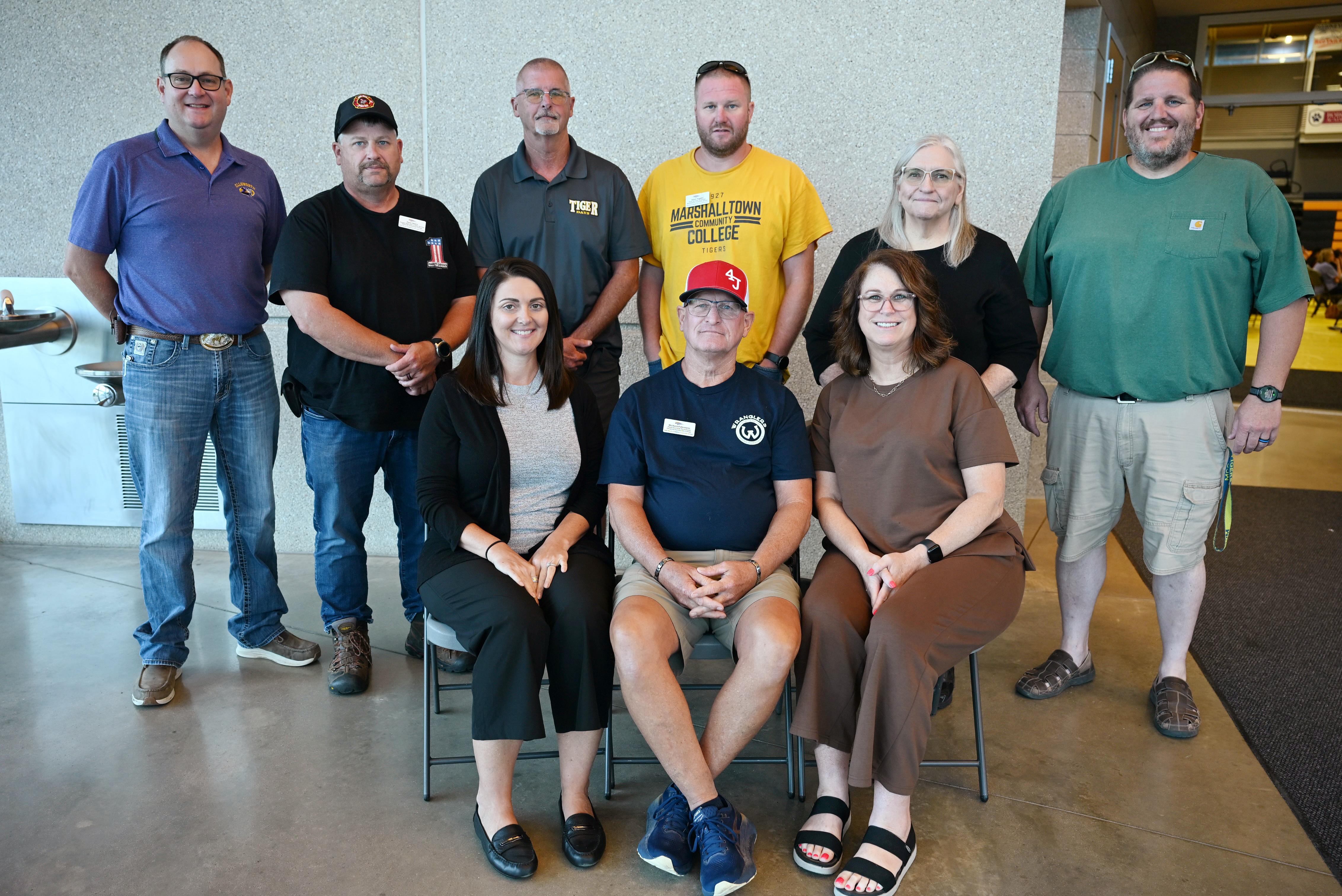 Nine employees pose for a group photo indoors, with three seated in the front row and six standing behind them against a textured wall during a workplace recognition event.