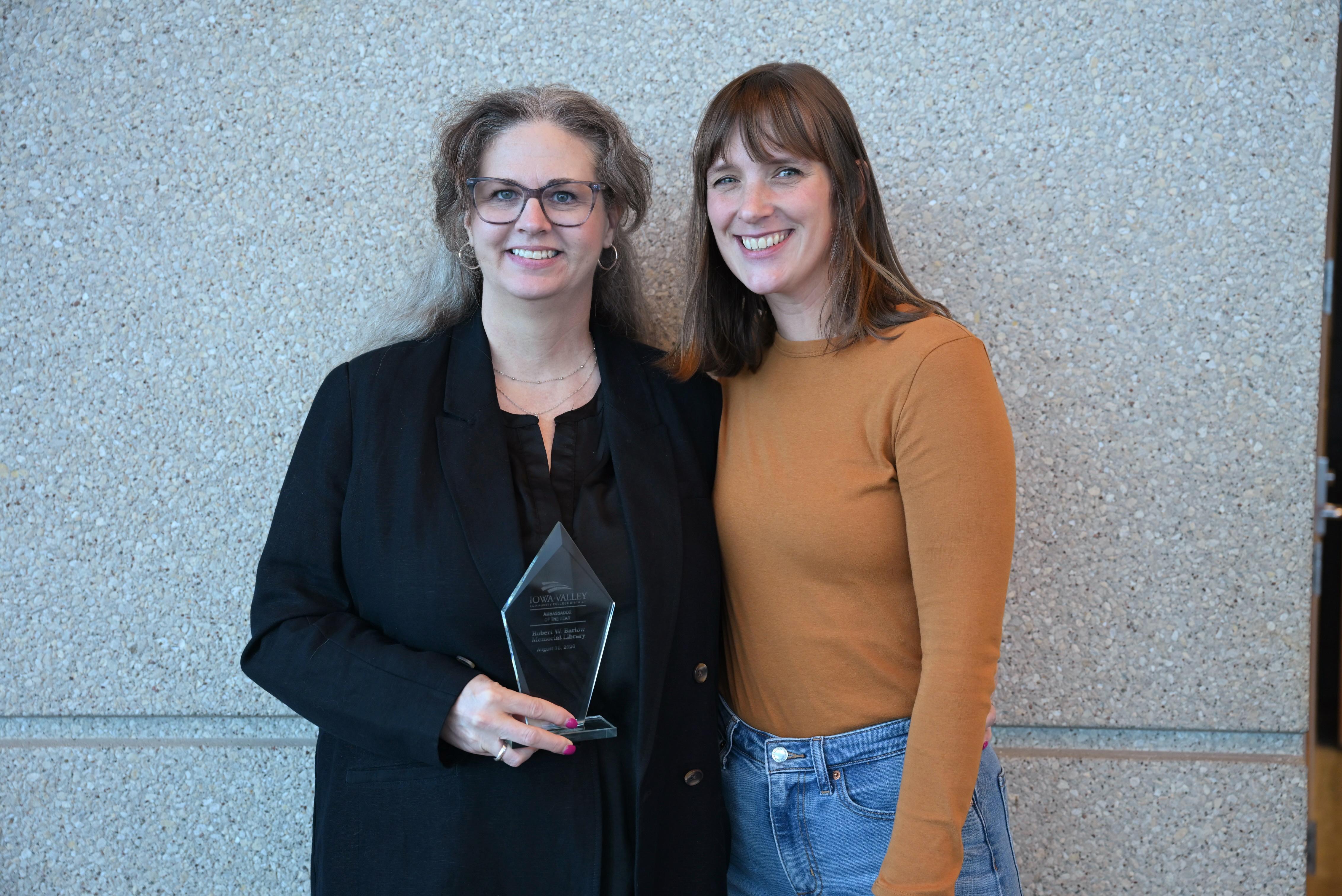 Two people pose together, with one holding a glass recognition award plaque.