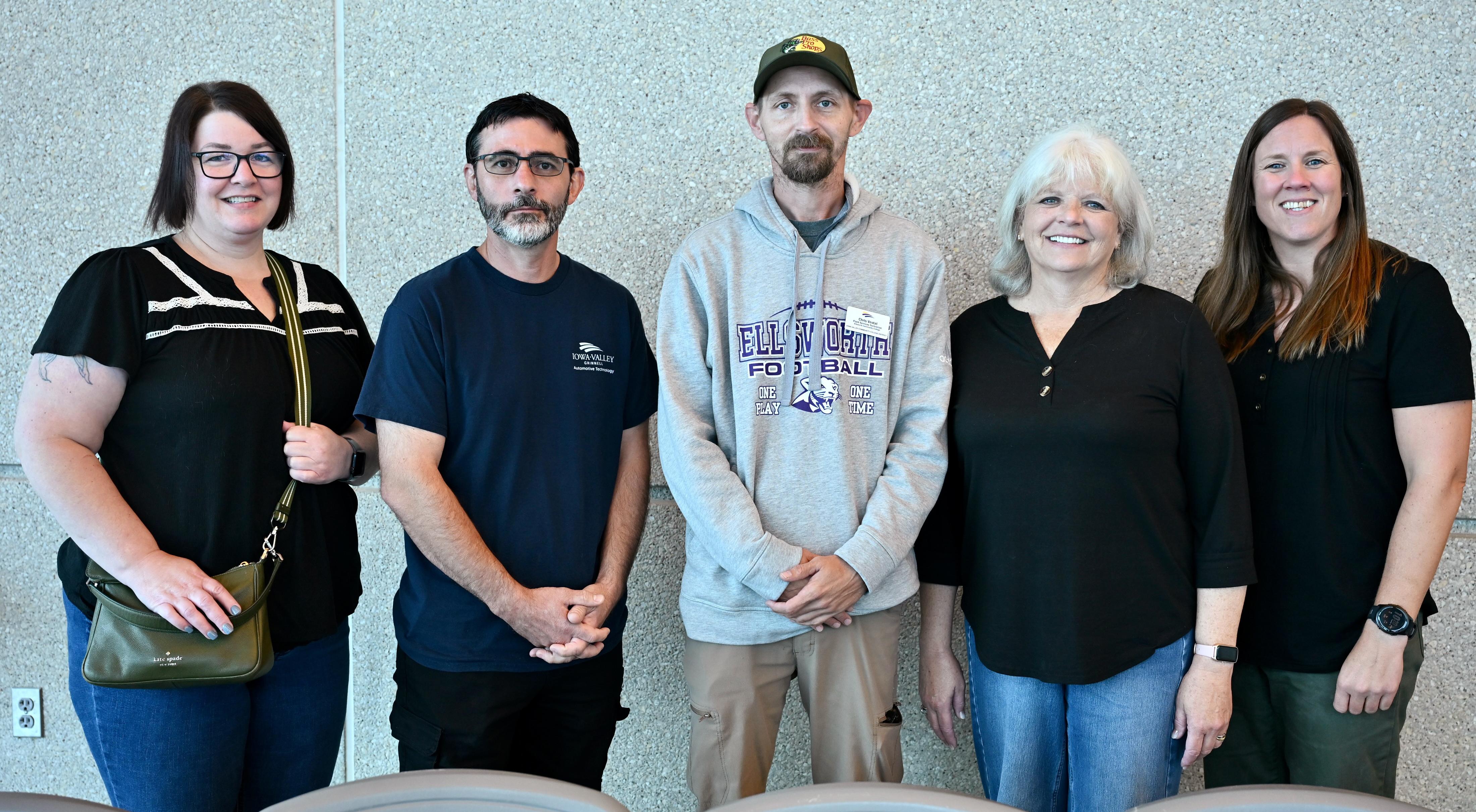 Five employees stand side by side in front of a textured indoor wall for a group recognition photo.