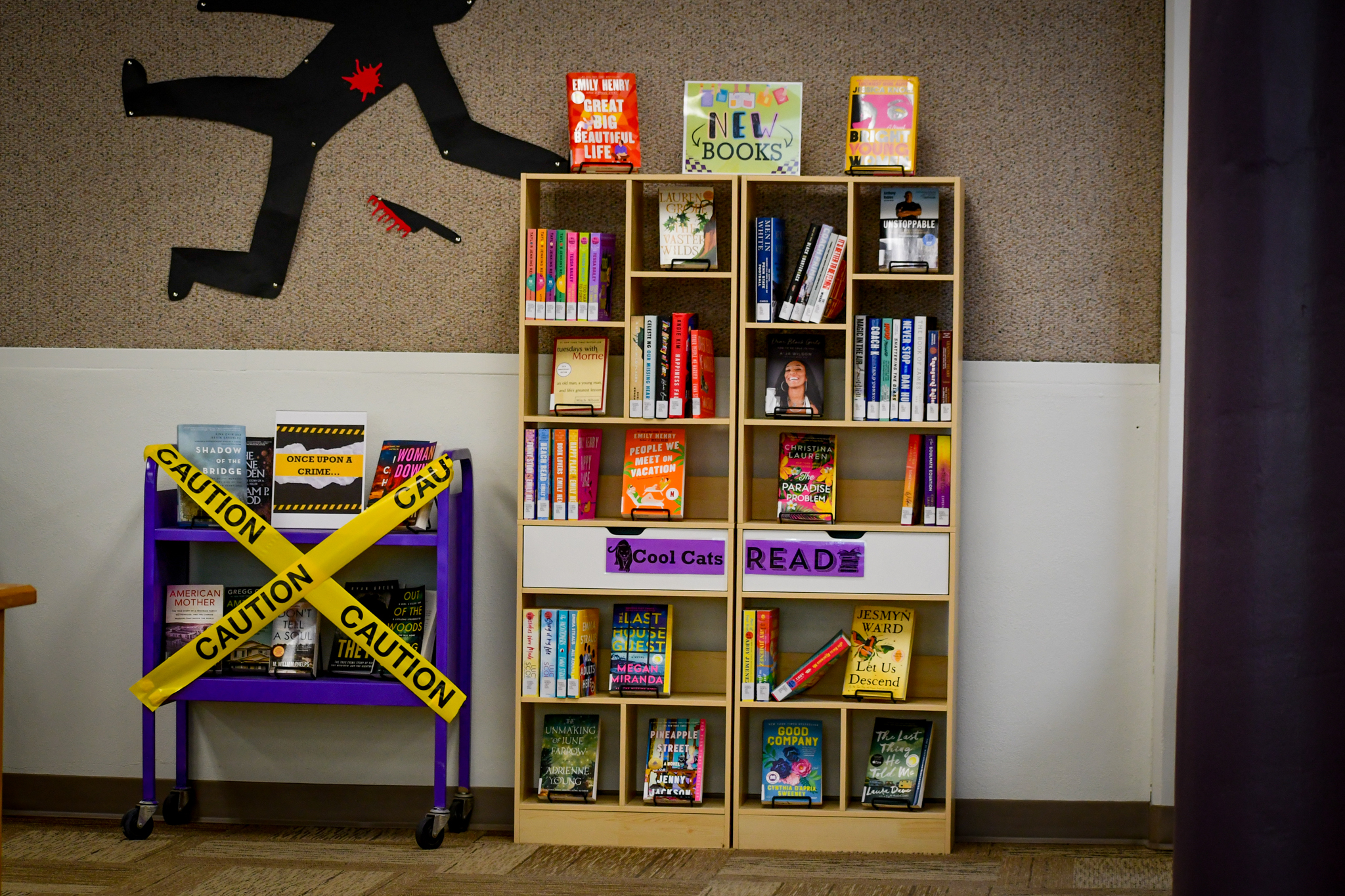 Halloween-themed library book display featuring shelves of new books, caution tape crossing a purple book cart, and a black silhouette with a red splatter decoration on the wall.