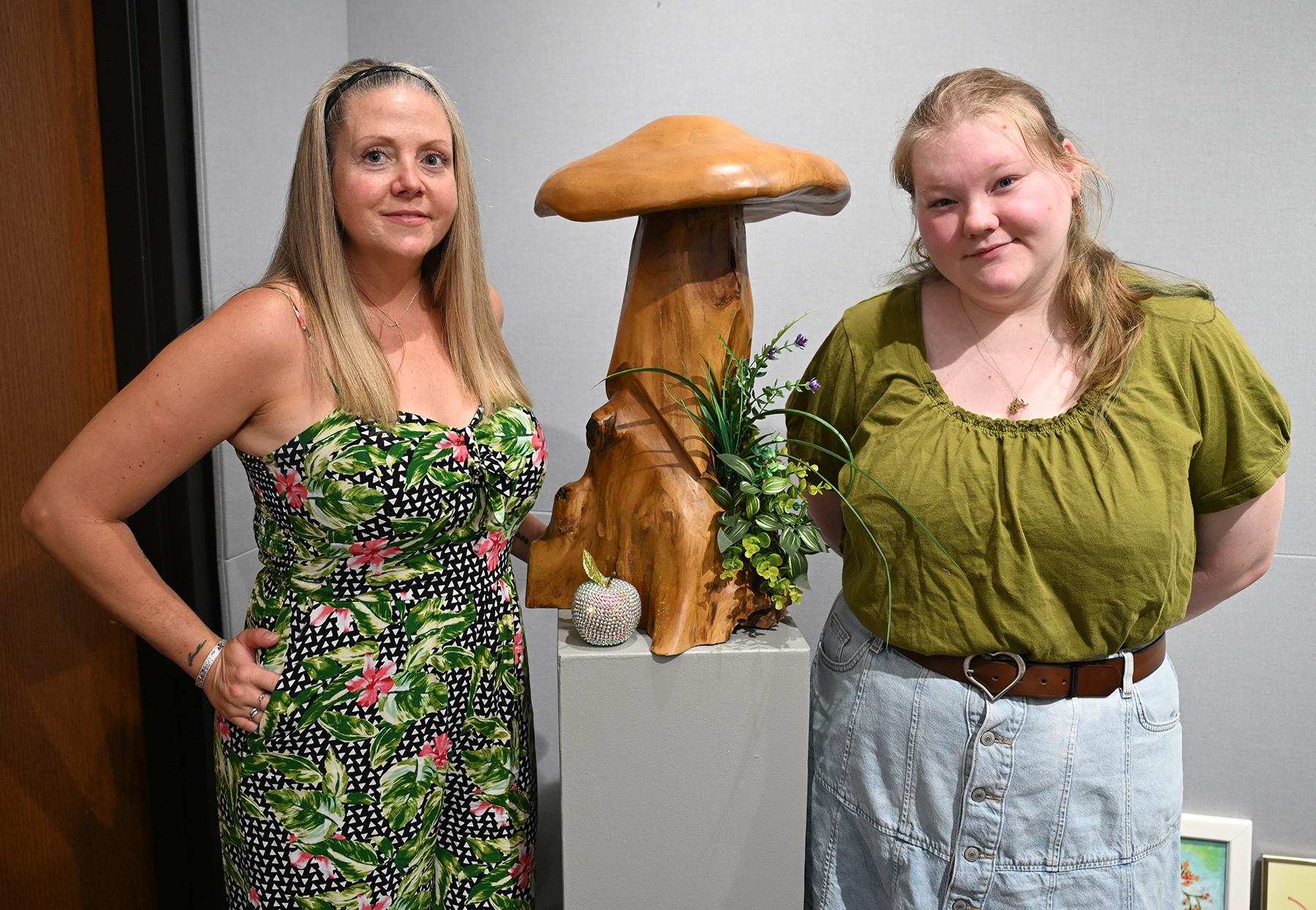 Two people stand on either side of a large polished wooden mushroom sculpture displayed on a pedestal indoors. One person wears a black floral-patterned dress, and the other wears a green blouse with a light denim skirt and brown belt. Decorative greenery and a small embellished pumpkin sit at the base of the sculpture against a light-colored wall.