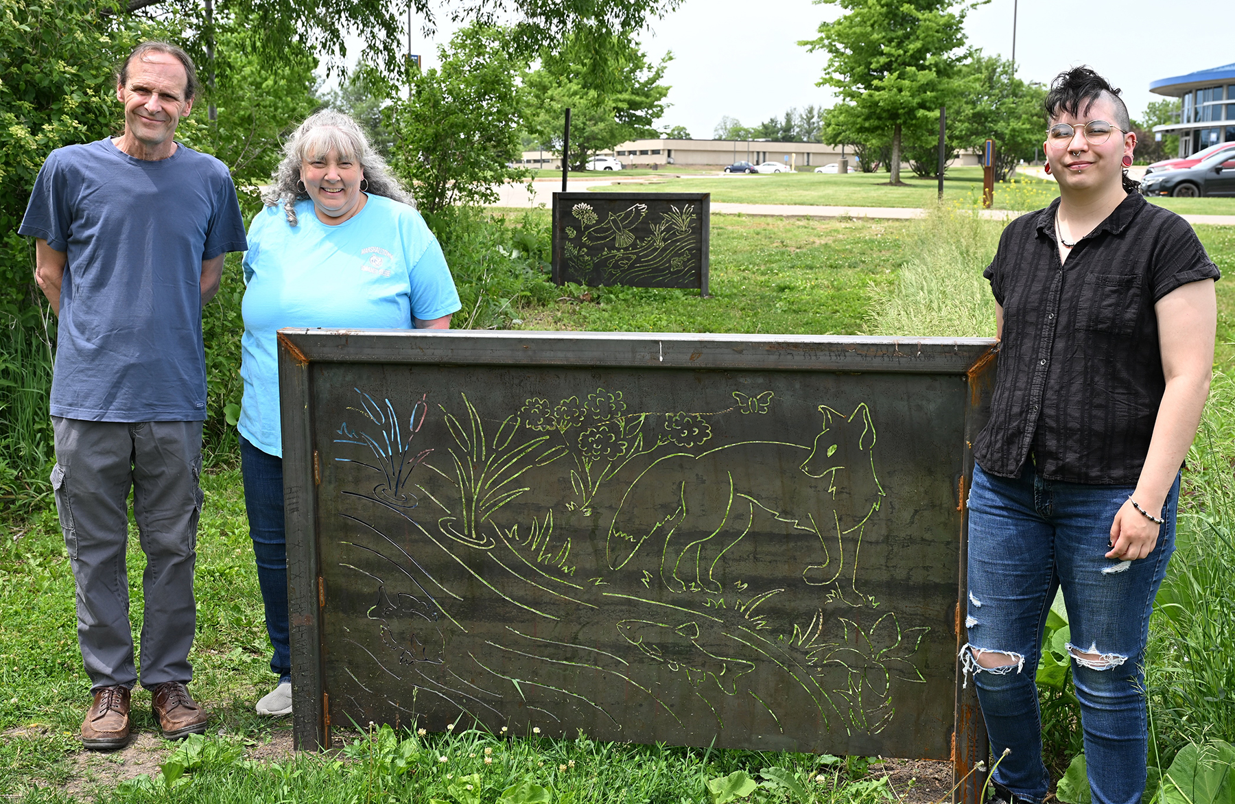 Three people stand outdoors beside a rectangular metal panel sculpture featuring cut-out designs of plants, water, fish, and a fox, installed in a grassy area with trees, a road, and campus buildings in the background.