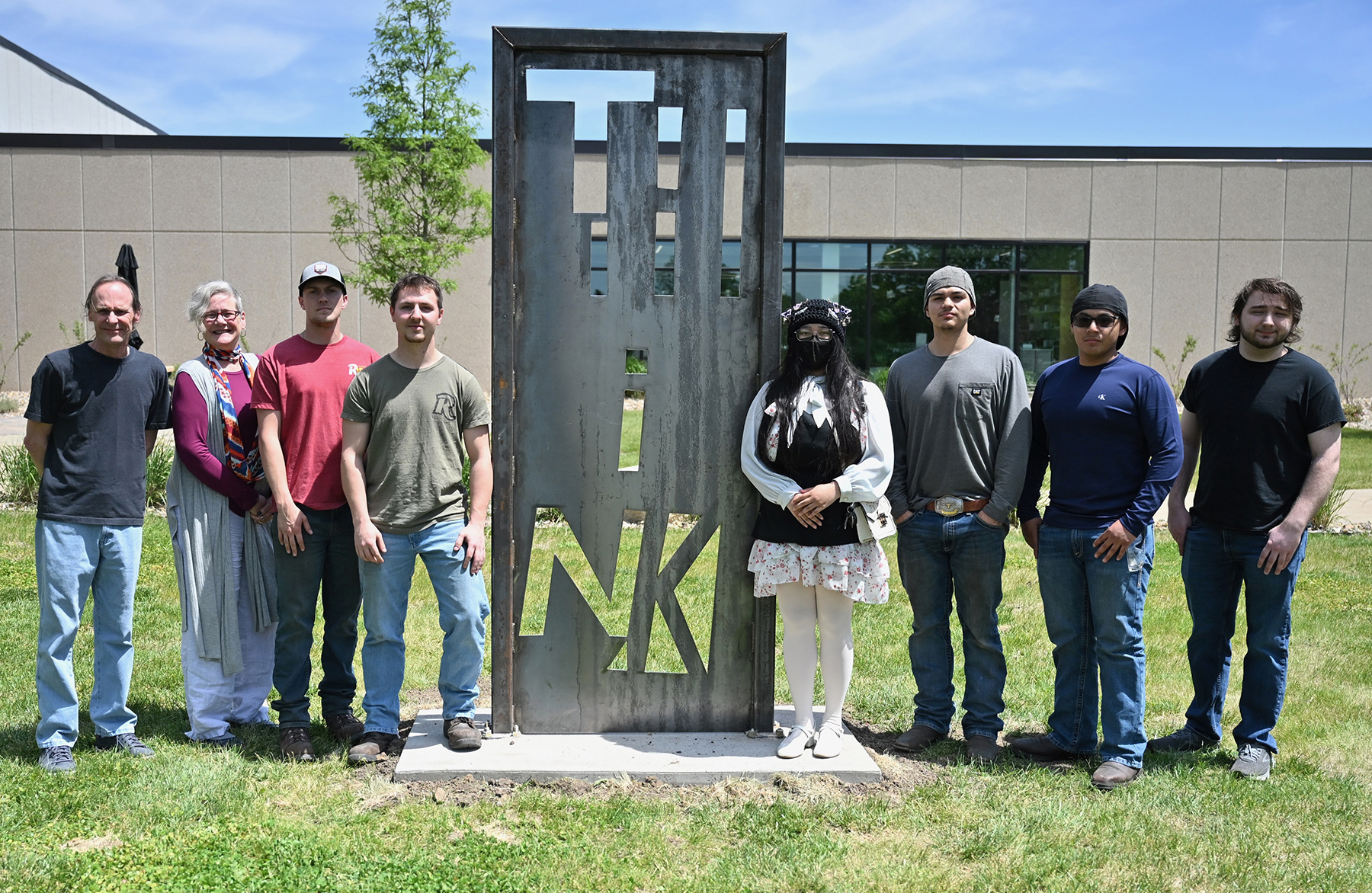 Group of eight people standing on a campus lawn around a tall, rectangular metal sculpture with cut-out letters, positioned in front of a college building and a small tree on a sunny day.